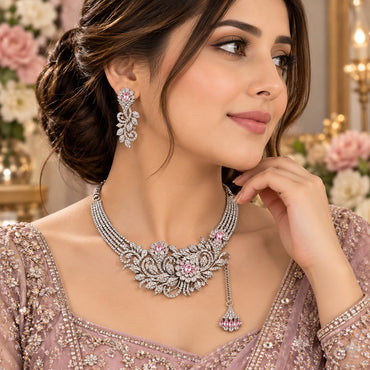 Silver White and Pink CZ Asymmetric Floral Vine Bib Necklace Earrings and Tikka Set worn by Pakistani female model in dusty rose lehenga in candlelit backdrop setting showing asymmetric floral vine bib necklace with dense white CZ micro-pave scrolling vine branches and oval pink CZ flower focal accents and matching long floral vine drop earrings