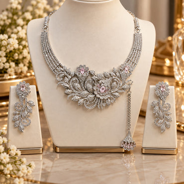 Silver White and Pink CZ Asymmetric Floral Vine Bib Necklace Earrings and Tikka Set displayed on white jewelry stand in luxury boutique setting with white gypsophila flowers and gold mirror backdrop showing 3-piece set with dramatic asymmetric floral vine bib necklace with white CZ tennis chain band and pink CZ flower accents with decorative side chain tassel drop and matching long floral vine drop earrings and tikka