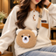 Tan curly-textured teddy-face bag with cream ears and a coordinating strap, worn crossbody over a cream sweatshirt.